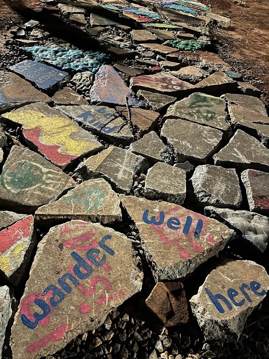 February 2023. Painted messages on the smashed-up bits of what was once an accessible paved path. "Be kind." "Wander well here."