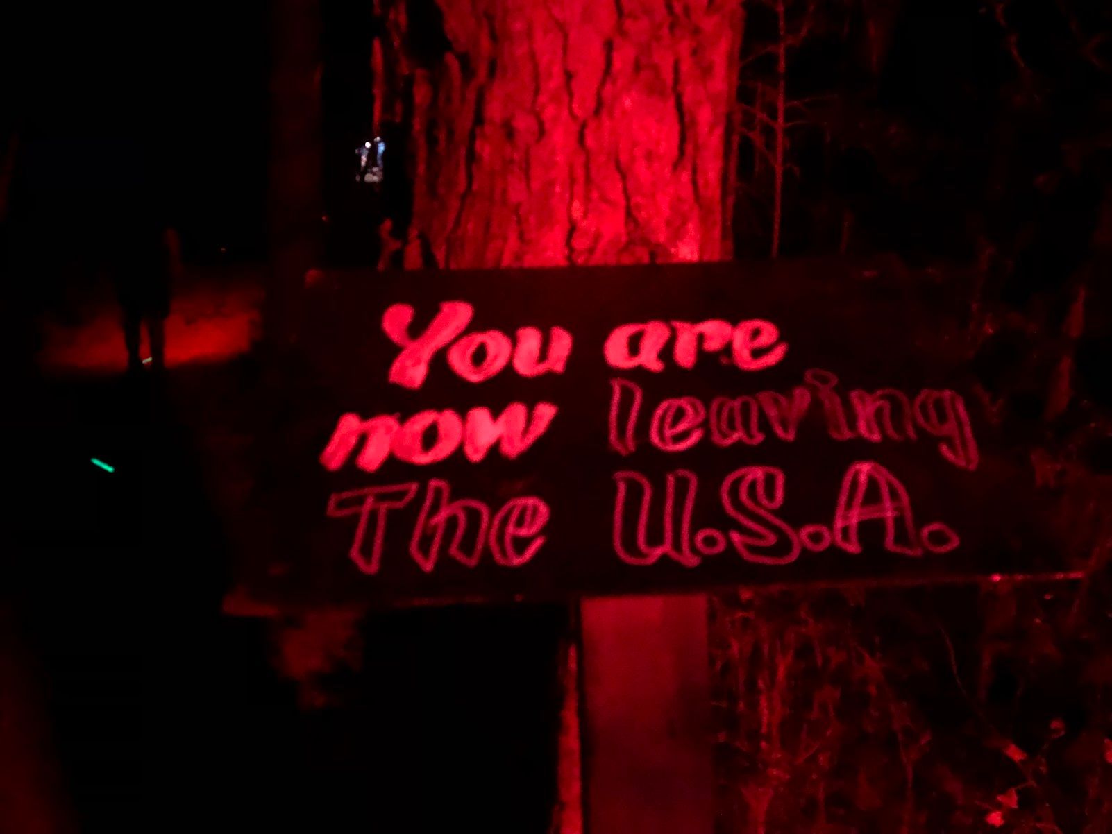 July 2022. A hand-painted sign reading "You are now leaving the U.S.A." The photo is awash in red light. Using red light at a nighttime campsite can help your eyes acclimate to the dark.